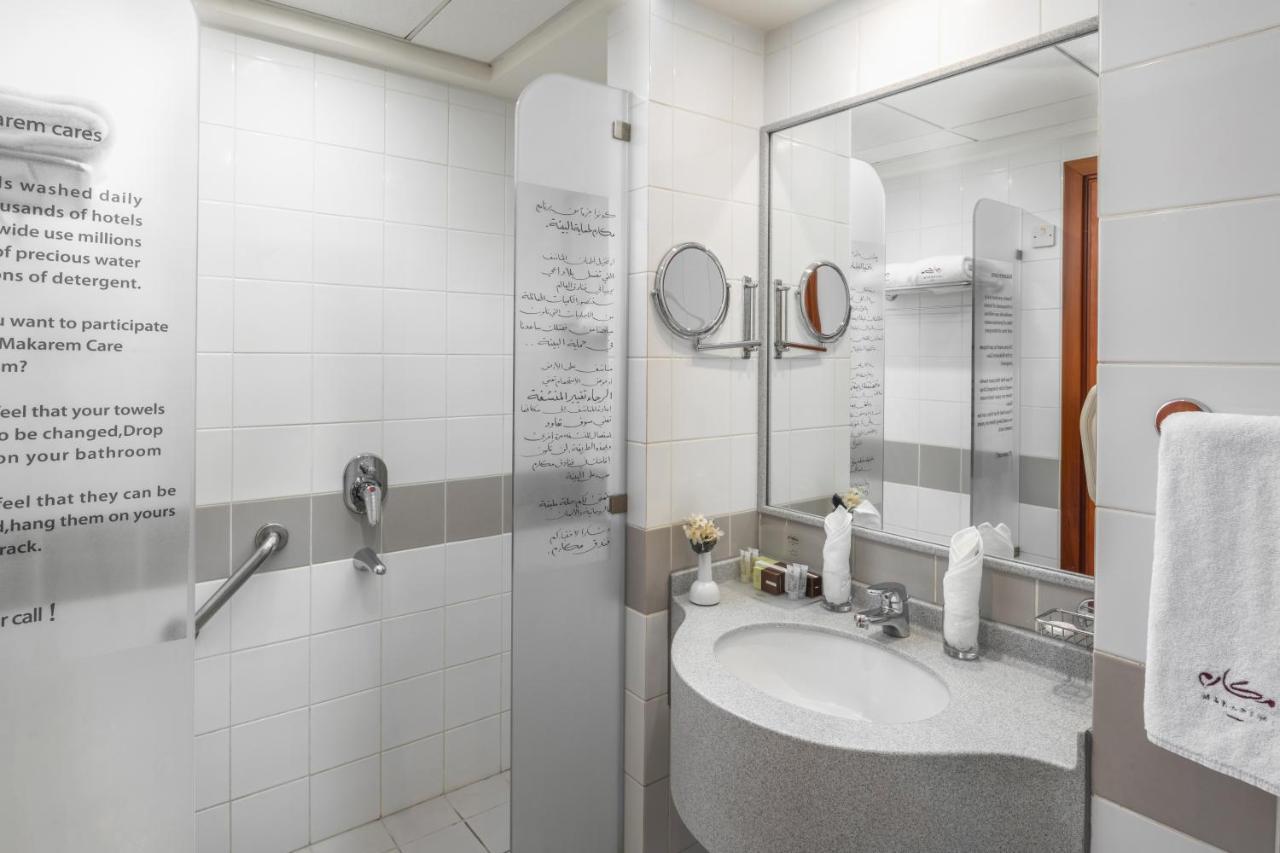 โรงแรมมากาเรม อุมม์ อัลกุรา Mecca ภายนอก รูปภาพ The photo shows a modern bathroom with a clean, minimalist design. It features a shower area separated by a glass panel, and there is a round mirror above a curved sink. The sink is accompanied by various toiletries and a hand towel. White tiles domi