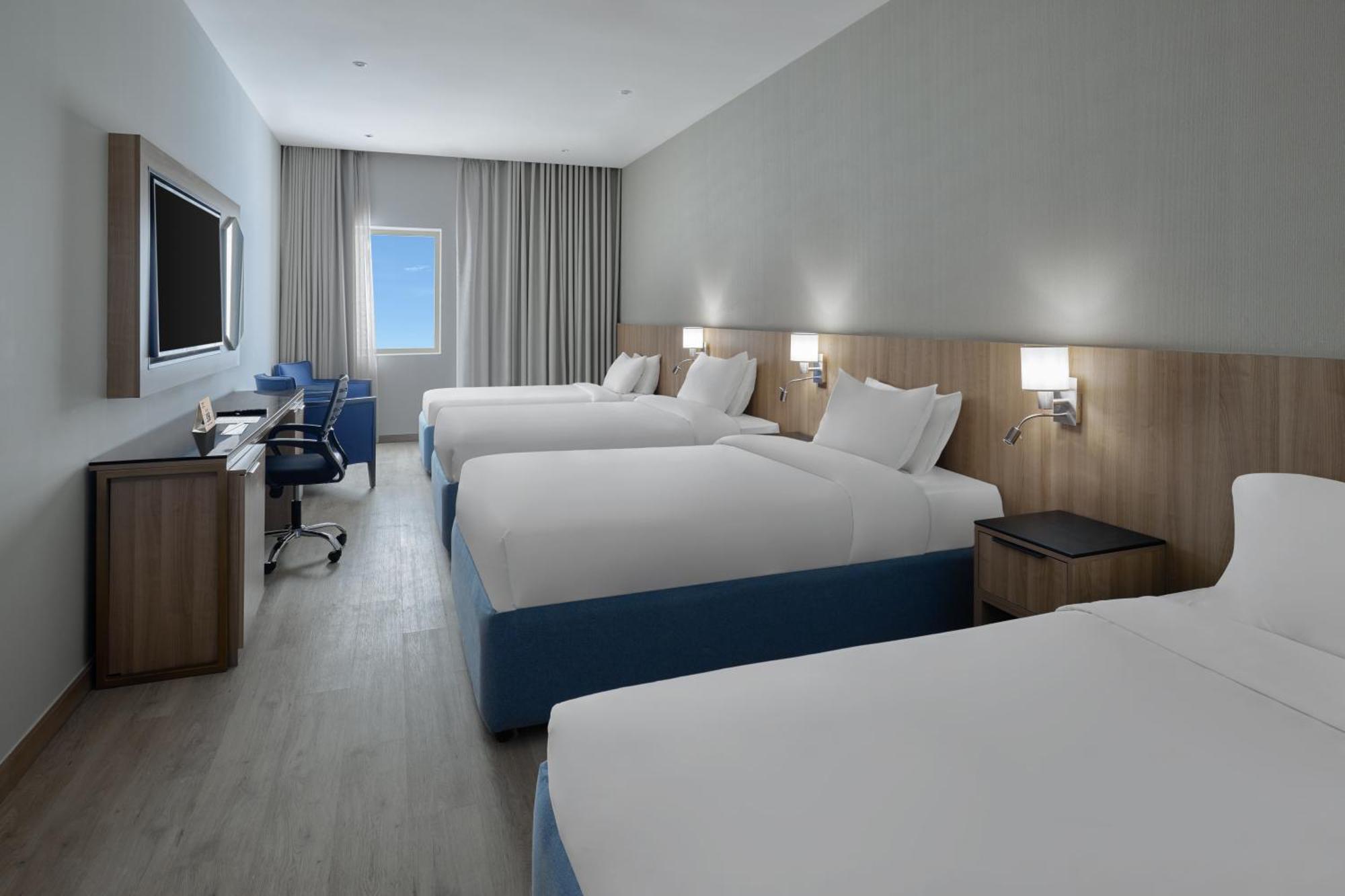 โรงแรมมากาเรม อุมม์ อัลกุรา Mecca ภายนอก รูปภาพ The photo shows a modern hotel room featuring four beds set up in a spacious layout. Each bed has white linens and blue accents. There is a large flat-screen TV mounted on the wall, and a desk with a chair is positioned next to it, providing a worksp