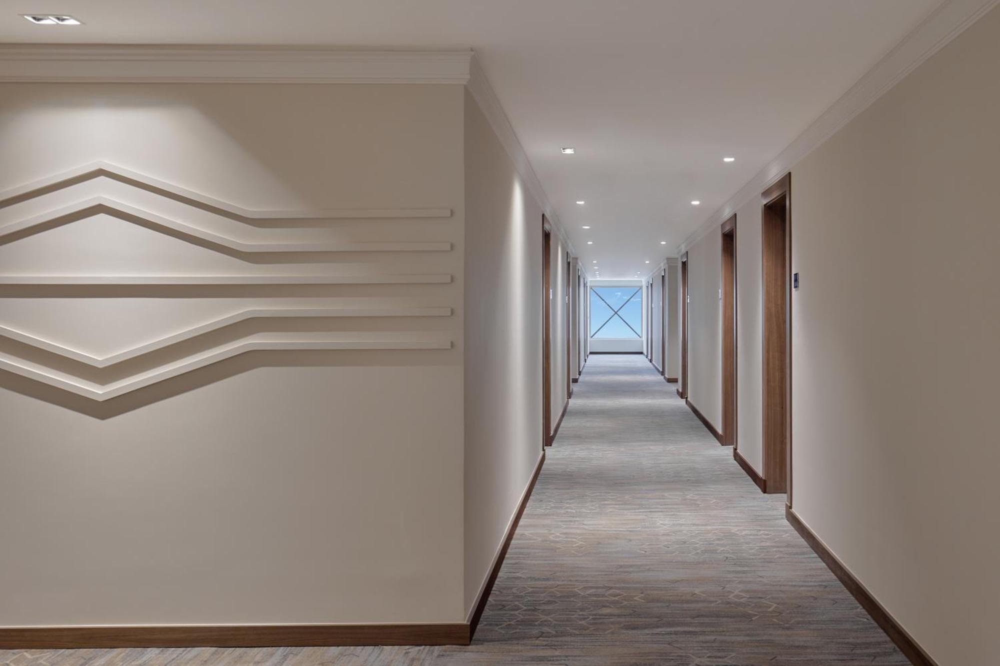 โรงแรมมากาเรม อุมม์ อัลกุรา Mecca ภายนอก รูปภาพ The photo shows a modern hotel corridor with a neutral color scheme. The walls are light-colored, and there are sleek, minimalistic features, including decorative lines on the left wall. The corridor itself is well-lit with recessed ceiling lights an