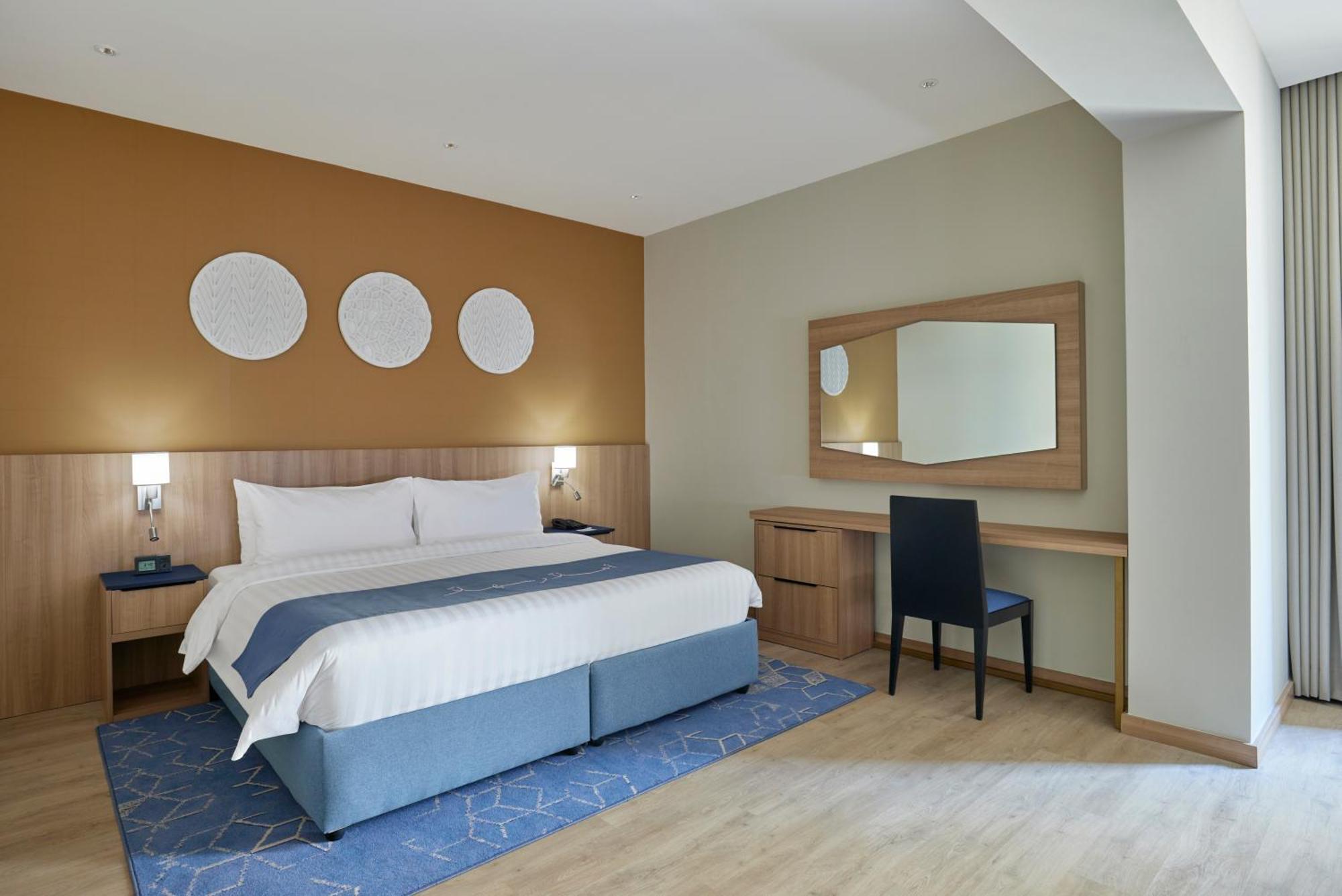 โรงแรมมากาเรม อุมม์ อัลกุรา Mecca ภายนอก รูปภาพ The photo shows a modern hotel room. It features a large bed with white linens and a blue base. On the wall above the bed are three circular decorative pieces. There is a desk with a mirror next to it, and a black chair is positioned in front of the 