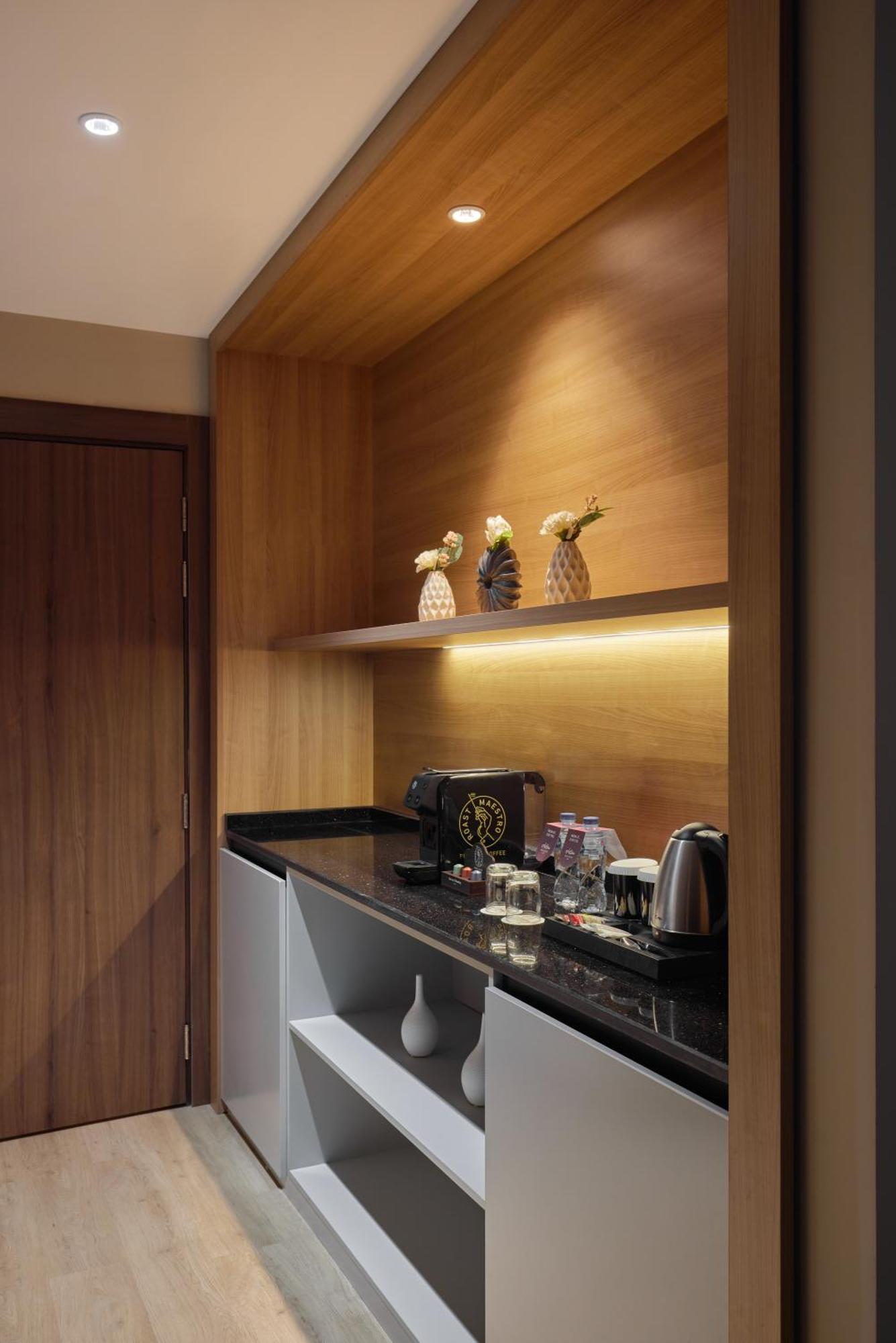 โรงแรมมากาเรม อุมม์ อัลกุรา Mecca ภายนอก รูปภาพ The photo shows a small kitchenette area with a wooden wall. There are three decorative vases with flowers on a shelf above a dark countertop. On the countertop, there is a coffee maker, a kettle, glasses, and bottles of water. The space is well-lit,
