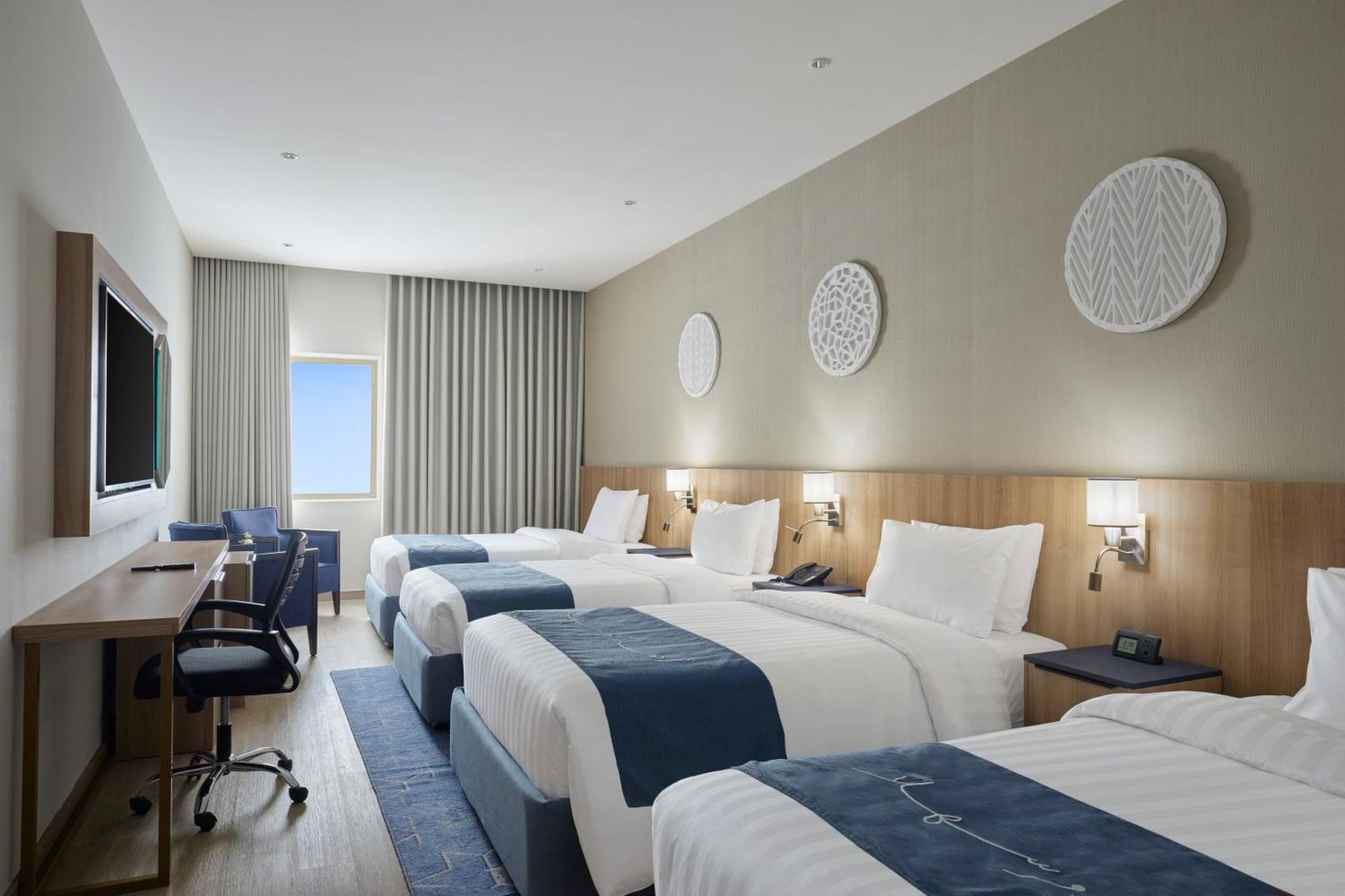 โรงแรมมากาเรม อุมม์ อัลกุรา Mecca ภายนอก รูปภาพ The photo shows a modern hotel room featuring three beds, all neatly made with white linens and blue accents. The room has soft, neutral-colored walls and wooden elements. There is a large television mounted on the wall opposite the beds and a work d