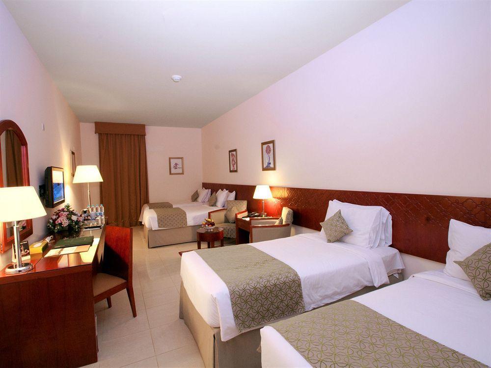 โรงแรมมากาเรม อุมม์ อัลกุรา Mecca ภายนอก รูปภาพ The photo shows a hotel room with a light, neutral color scheme. There are two twin beds on one side, each with white linens and decorative throw pillows. A bedside table with a lamp is positioned between the beds. On the other side of the room, ther