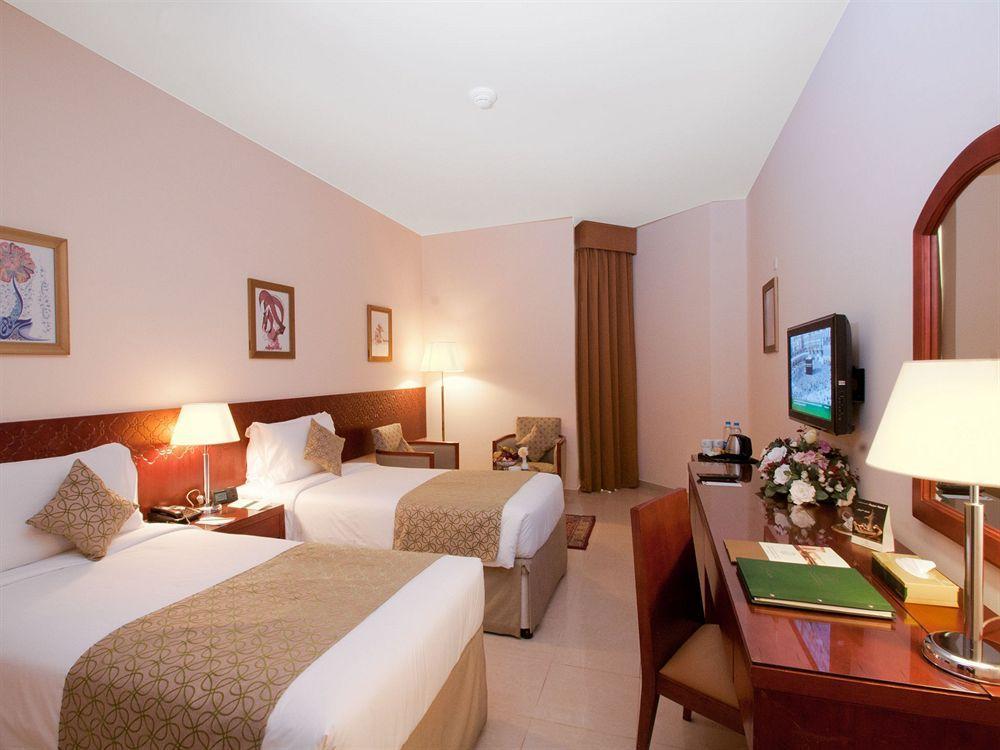โรงแรมมากาเรม อุมม์ อัลกุรา Mecca ภายนอก รูปภาพ The photo shows a hotel room featuring two single beds with light-colored bedding and decorative throw pillows. There are bedside tables with lamps next to each bed, and wall art is displayed above the beds. A chair is positioned near the window, and