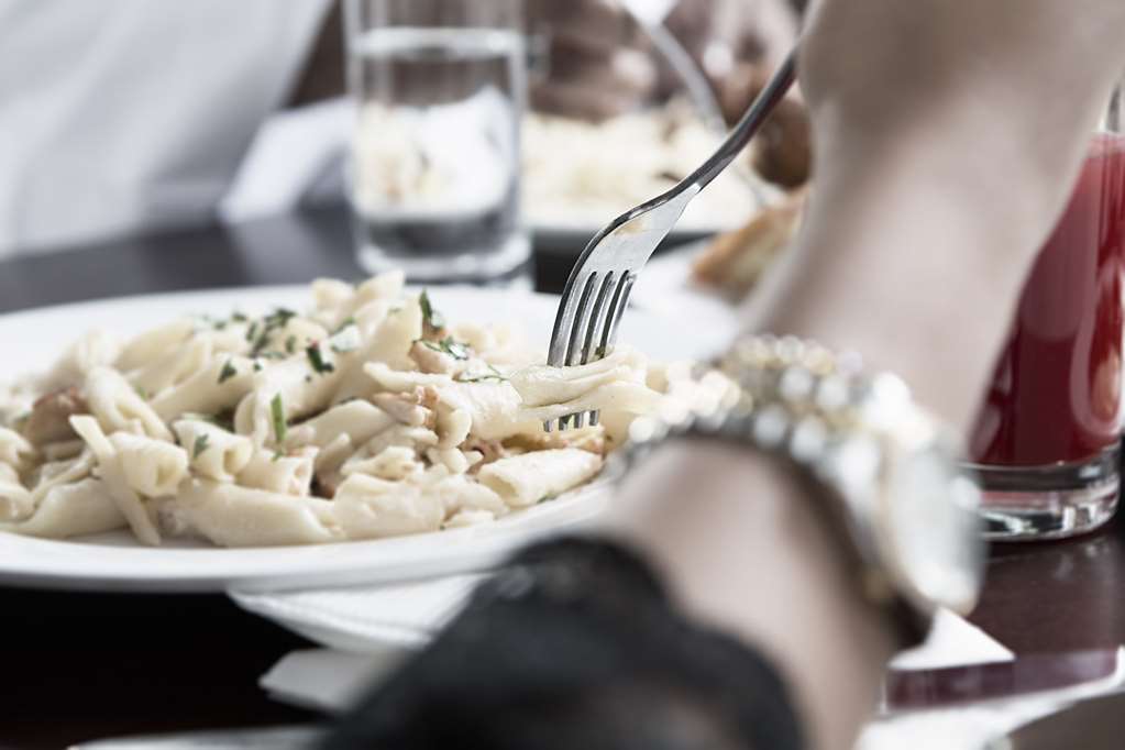 โรงแรมมากาเรม อุมม์ อัลกุรา Mecca ร้านอาหาร รูปภาพ The photo shows a close-up of a plate of pasta garnished with herbs, with a fork poised above it, suggesting someone is about to take a bite. To the side, there is a hand wearing a watch, and in the background, there are glasses of water on the table