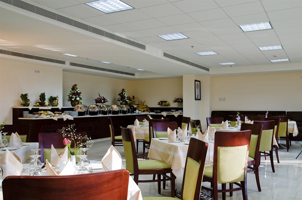 โรงแรมมากาเรม อุมม์ อัลกุรา Mecca ภายนอก รูปภาพ The photo shows a dining area in a restaurant or banquet hall. There are several tables arranged with cream-colored tablecloths and neatly folded napkins. The chairs have a mix of dark wood and light green upholstery. Along the back wall, there is a 