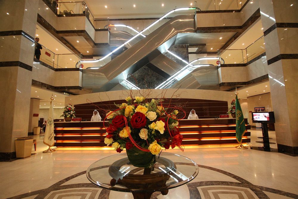 โรงแรมมากาเรม อุมม์ อัลกุรา Mecca ภายนอก รูปภาพ The photo shows a modern hotel lobby. In the foreground, there is an elegant floral arrangement featuring various colorful flowers in a decorative vase. In the background, a reception desk is visible, staffed by individuals in uniform. Two escalators
