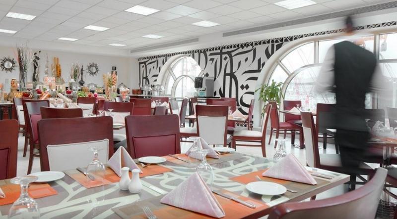 โรงแรมมากาเรม อุมม์ อัลกุรา Mecca ภายนอก รูปภาพ The photo shows the interior of a restaurant. It features a modern design with a combination of wooden and white furniture. The tables are set with white plates and folded napkins, arranged neatly. There are decorative touches such as small plants an