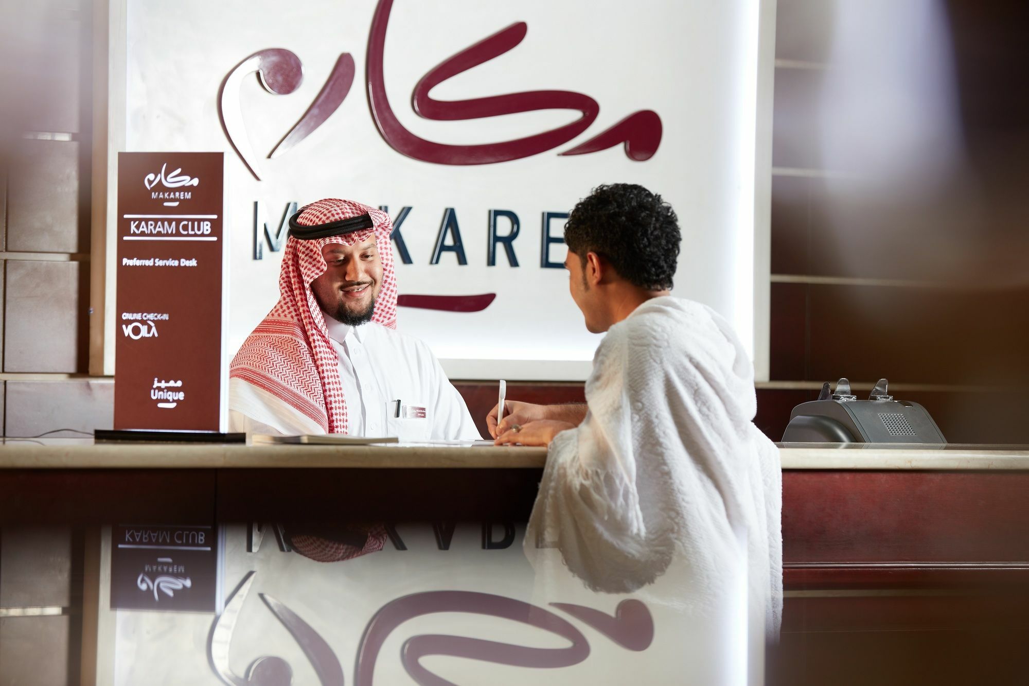 โรงแรมมากาเรม อุมม์ อัลกุรา Mecca ภายนอก รูปภาพ The photo shows a scene at a reception area. There are two men, one dressed in traditional Saudi attire with a red and white keffiyeh, who appears to be a staff member at a service counter. The other man, wearing a white robe, is engaged in conversat