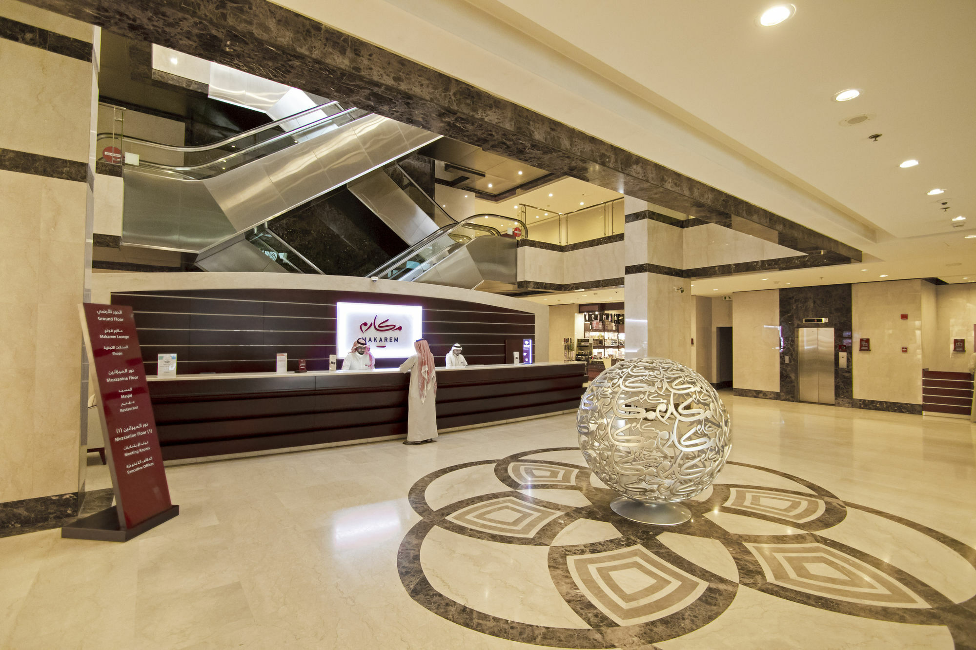 โรงแรมมากาเรม อุมม์ อัลกุรา Mecca ภายนอก รูปภาพ The photo shows the interior of a hotel lobby. There is a reception desk with staff attending to guests, and a modern, elegant design is evident throughout the space. In the center, there is a distinctive decorative spherical piece, likely made of gl